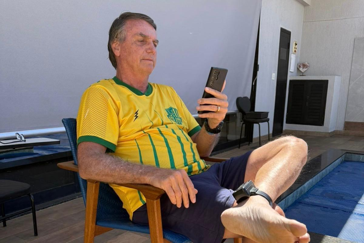 ex-presidente Jair Bolsonaro (PL) sentado, com tornozeleira e pernas cruzadas, de camiseta da CBF