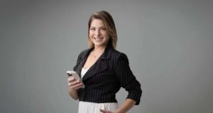 A jornalista Daniela Lima de cabelo solto, sorrindo para a câmera, de roupa social, segurando celular