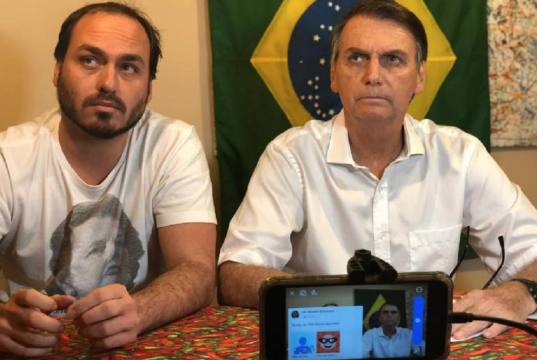 O vereador Carlos Bolsonaro (PL-RJ) e o ex-presidente Jair Bolsonaro (PL) sentados lado a lado, sérios, sem olhar para a câmera