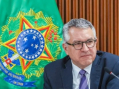 O ministro da Saúde, Alexandre Padilha, sério, olhando para o lado, de óculos, com bandeira ao fundo