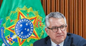 O ministro da Saúde, Alexandre Padilha, sério, olhando para o lado, de óculos, com bandeira ao fundo