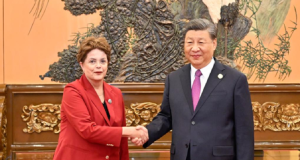 Dilma Rousseff e o presidente Xi Jinping em aperto de mãos, olhando para a câmera