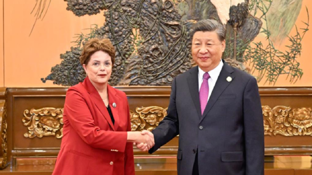Dilma Rousseff e o presidente Xi Jinping em aperto de mãos, olhando para a câmera
