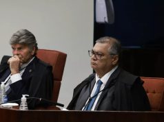 Luiz Fux e Flávio Dino sérios, sentados lado a lado, sem olhar para a câmera