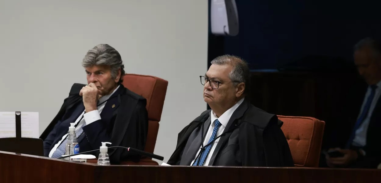 Luiz Fux e Flávio Dino sérios, sentados lado a lado, sem olhar para a câmera