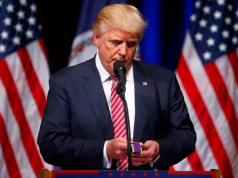 Donald Trump olhando para baixo, sério, perto de microfone, em pé