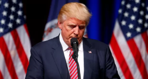 Donald Trump olhando para baixo, sério, perto de microfone, em pé