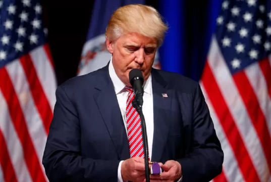 Donald Trump olhando para baixo, sério, perto de microfone, em pé