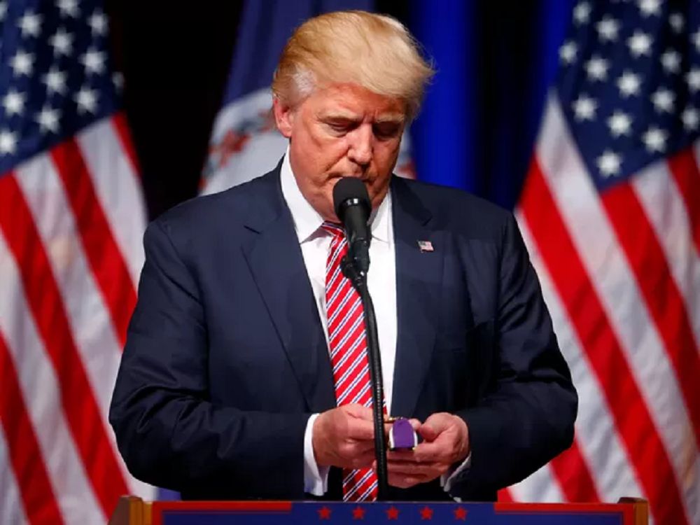 Donald Trump olhando para baixo, sério, perto de microfone, em pé
