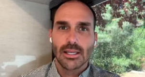 O deputado federal Eduardo Bolsonaro (PL-SP) em close, sério, falando para a câmera, usando fones de ouvido brancos sem fio