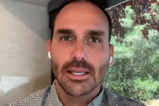 O deputado federal Eduardo Bolsonaro (PL-SP) em close, sério, falando para a câmera, usando fones de ouvido brancos sem fio