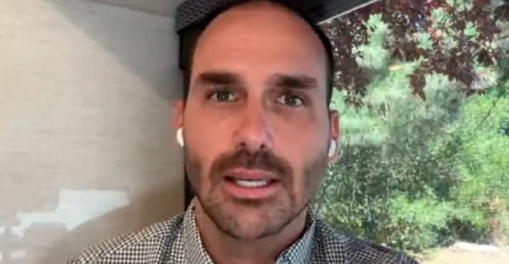 O deputado federal Eduardo Bolsonaro (PL-SP) em close, sério, falando para a câmera, usando fones de ouvido brancos sem fio