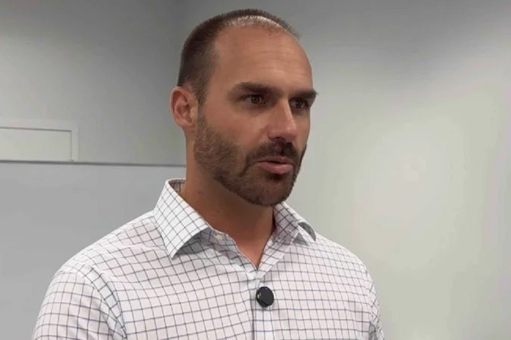 Eduardo Bolsonaro falando, sério, de camisa polo clara e quadriculada, sem olhar para a câmera