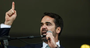 O governador Eduardo Leite (PSD) falando em microfone e apontando para o lado, sério