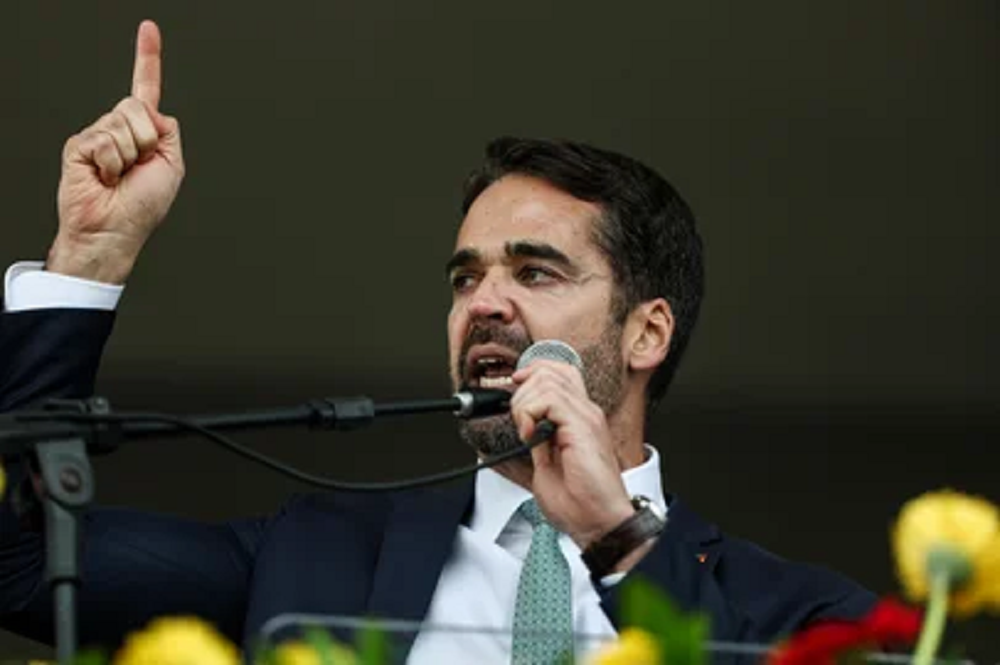 O governador Eduardo Leite (PSD) falando em microfone e apontando para o lado, sério