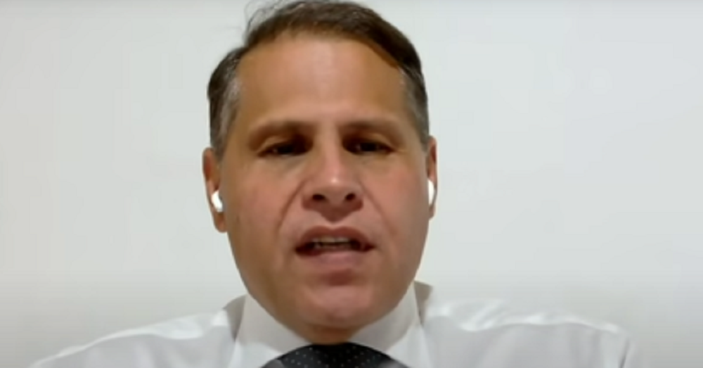 Eduardo Tagliaferro em close, de roupa social branca e fones de ouvido sem fio brancos, falando para a câmera