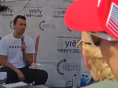 Charlie Kirk momentos antes de ser baleado, de calça escura e camiseta branca, falando em microfone, sentado