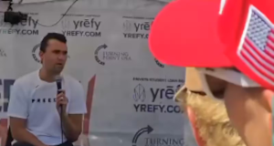 Charlie Kirk momentos antes de ser baleado, de calça escura e camiseta branca, falando em microfone, sentado