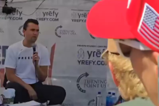 Charlie Kirk momentos antes de ser baleado, de calça escura e camiseta branca, falando em microfone, sentado