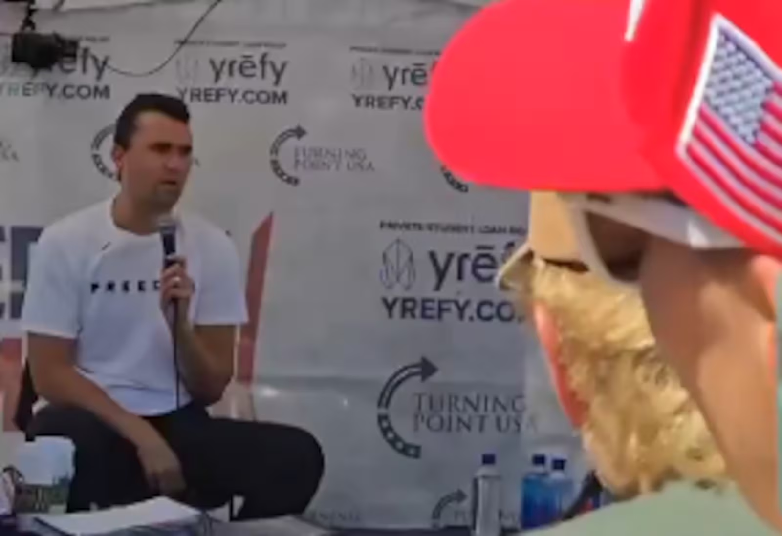 Charlie Kirk momentos antes de ser baleado, de calça escura e camiseta branca, falando em microfone, sentado