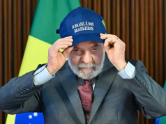 Lula sério, olhando pra frente, colocando boné, sentado, com bandeira do Brasil ao fundo