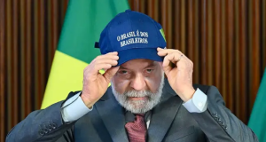 Lula sério, olhando pra frente, colocando boné, sentado, com bandeira do Brasil ao fundo