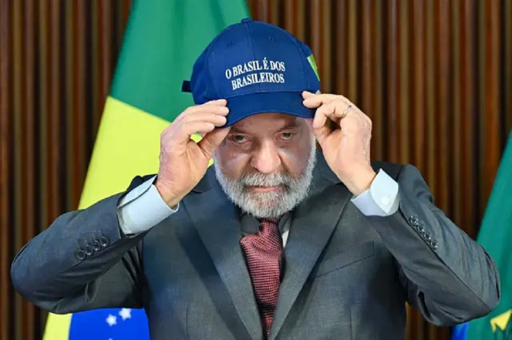 Lula sério, olhando pra frente, colocando boné, sentado, com bandeira do Brasil ao fundo