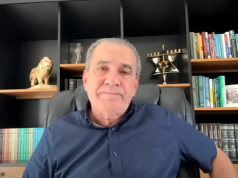 O pastor Silas Malafaia de roupa azul, olhando sério para a câmera, sentado na frente de estante preta cheia de objetos