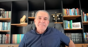 O pastor Silas Malafaia de roupa azul, olhando sério para a câmera, sentado na frente de estante preta cheia de objetos