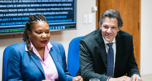 Margareth Menezes (Cultura) e Fernando Haddad (Economia) sentados lado a lado, ele sorrindo e ela séria
