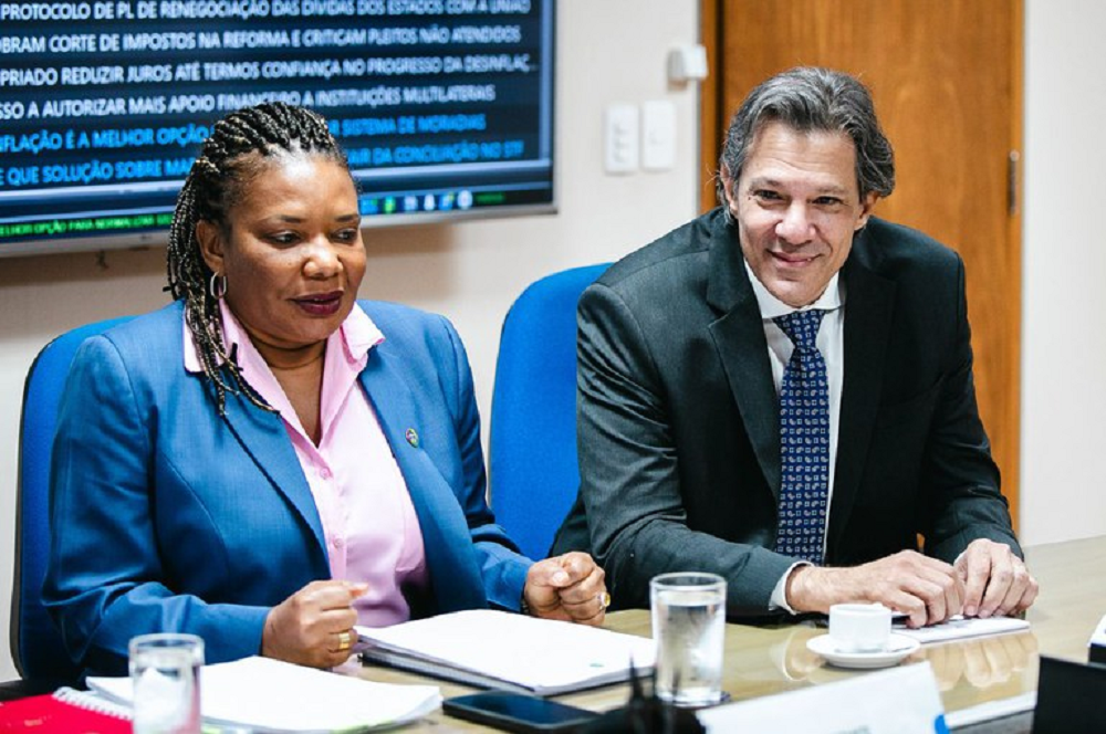 Margareth Menezes (Cultura) e Fernando Haddad (Economia) sentados lado a lado, ele sorrindo e ela séria