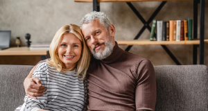 Casal de meia idade sentado no sofá, abraçado, sorrindo para a câmera