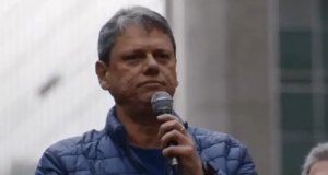Tarcísio de Freitas de roupa de frio azul, sério, segurando microfone