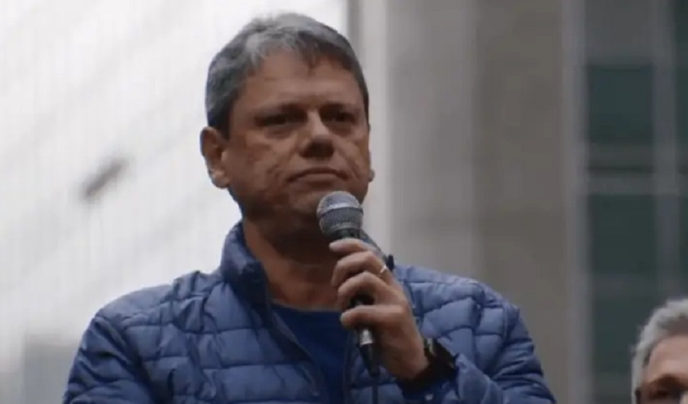Tarcísio de Freitas de roupa de frio azul, sério, segurando microfone