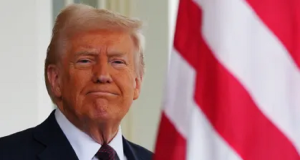 O presidente dos Estados Unidos, Donald Trump, em close, sorrindo de maneira debochada, com bandeira dos EUA ao fundo