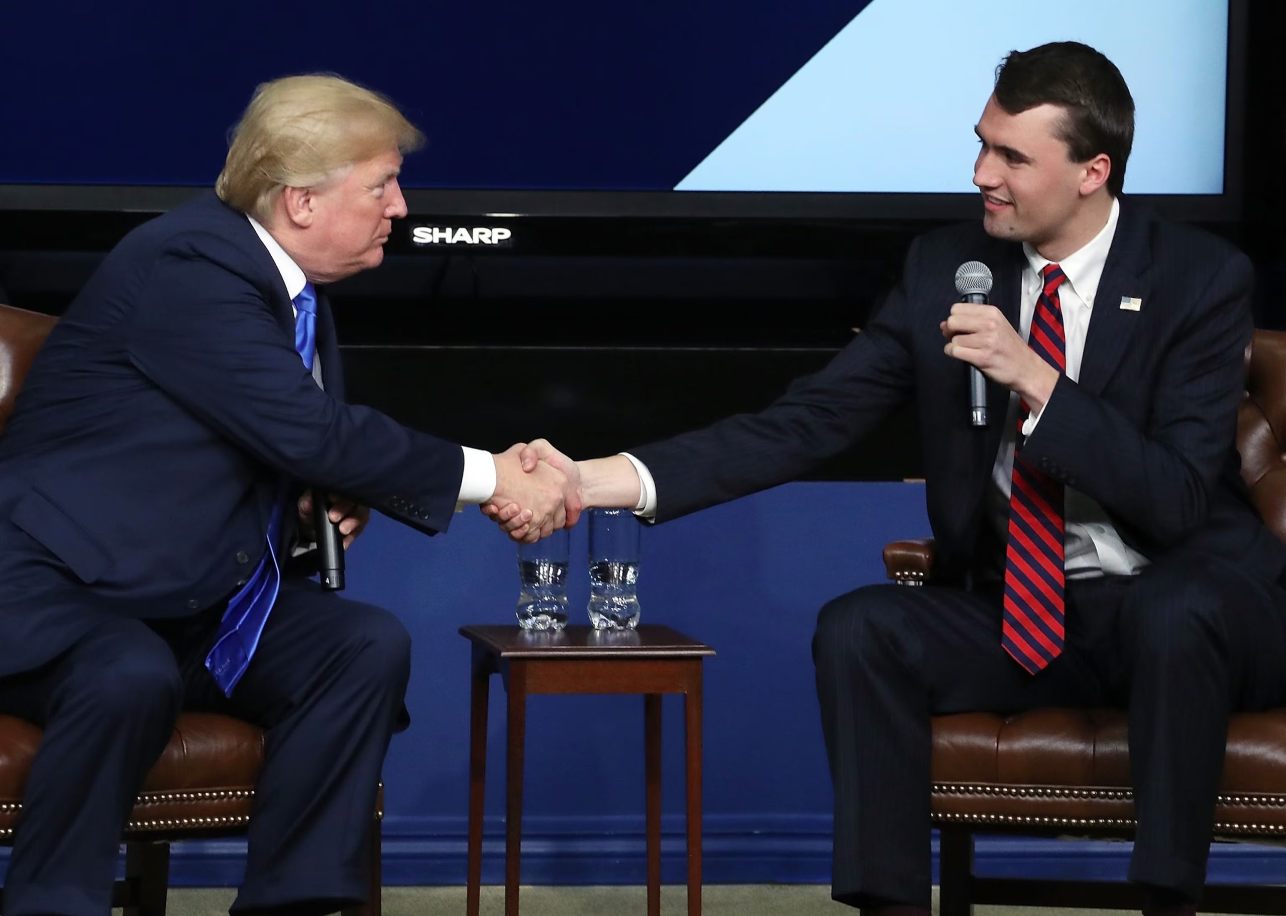O presidente dos EUA, Donald Trump, e o ativista conservador Charlie Kirk em aperto de mãos, se olhando, sentados em cadeiras próximas