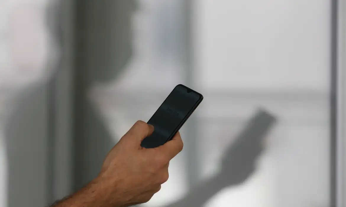 Mão mexendo no celular com sombra de homem ao fundo