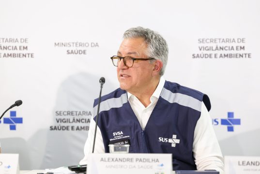 O ministro da Saúde, Alexandre Padilha, falando em microfone sem olhar para a câmera, de óculos
