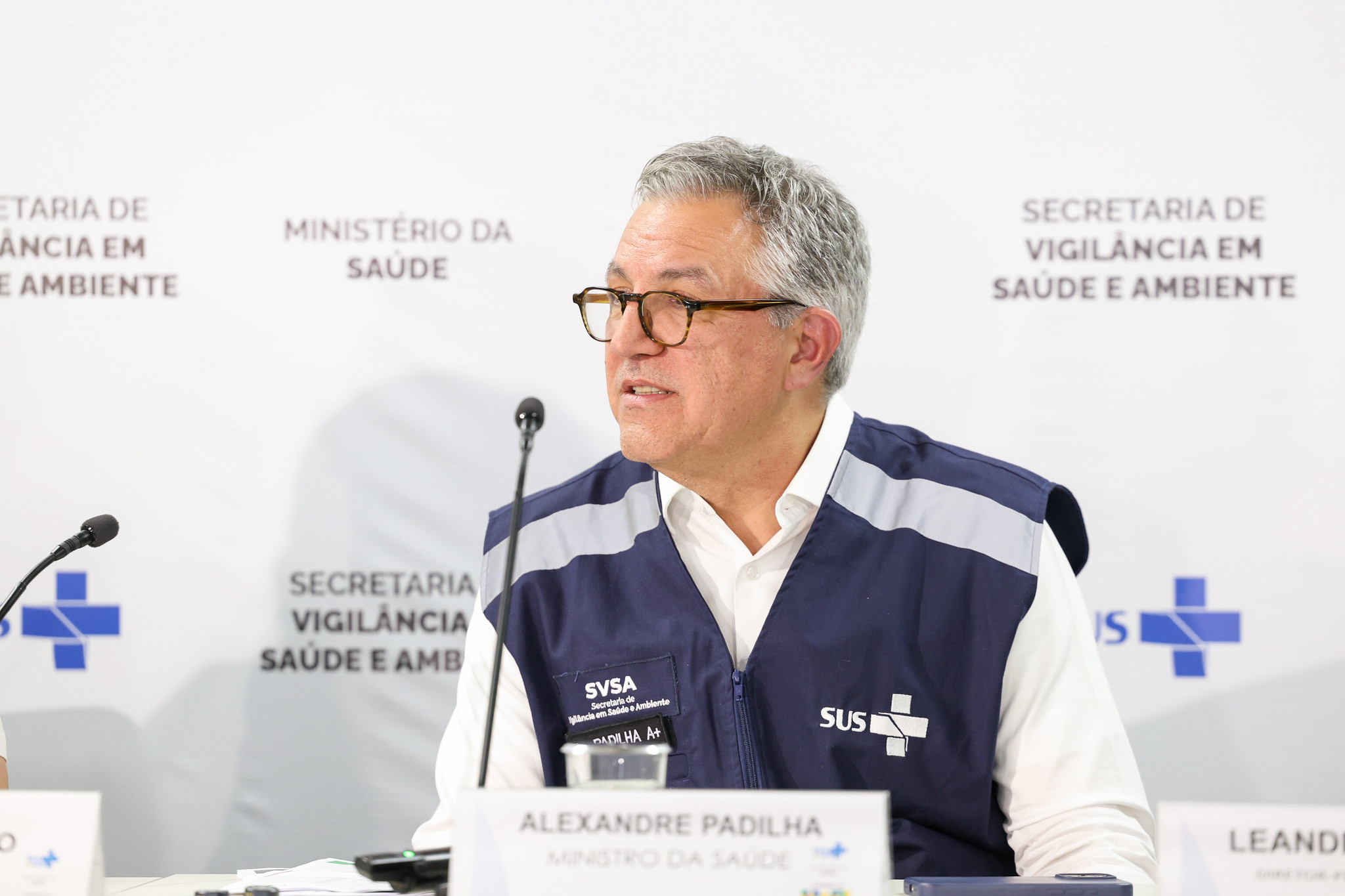 O ministro da Saúde, Alexandre Padilha, falando em microfone sem olhar para a câmera, de óculos