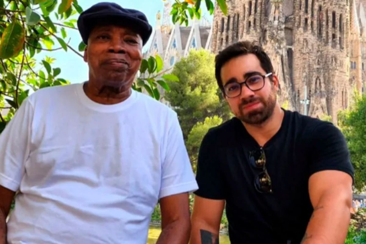 Milton Nascimento de boina preta e camiseta branca posando sentado ao lado de Augusto Nascimento, que veste camisa preta e óculos de grau
