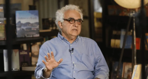 O escritor brasileiro Milton Hatoum de camisa social azul e óculos, falando e gesticulando sem olhar para a câmera, sério, sentado