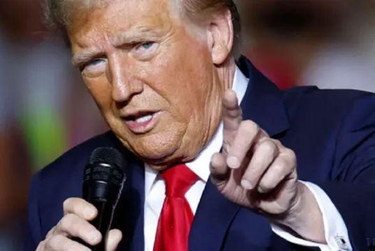 Donald Trump, presidente dos EUA, falando em microfone e apontando pra frente, em close