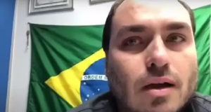 O vereador Carlos Bolsonaro (PL-RJ) no canto direito de foto, com cara de choro e bandeira do Brasil ao fundo
