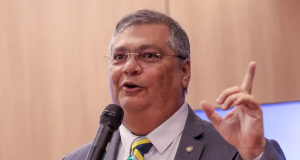 O ministro Flávio Dino, presidente da Primeira Turma do Supremo Tribunal Federal (STF), falando em microfone, sorrindo e apontando pra cima