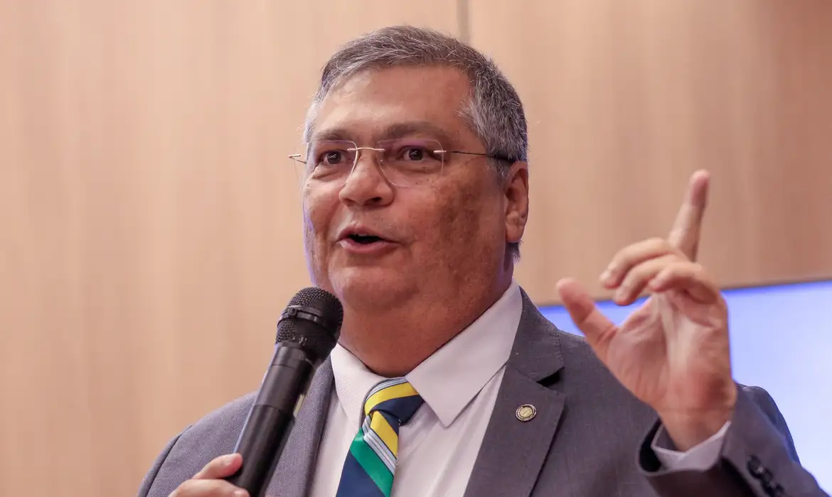 O ministro Flávio Dino, presidente da Primeira Turma do Supremo Tribunal Federal (STF), falando em microfone, sorrindo e apontando pra cima