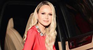 Eliana de vestido vermelho e cabelo solto, séria, olhando para a câmera, saindo de carro