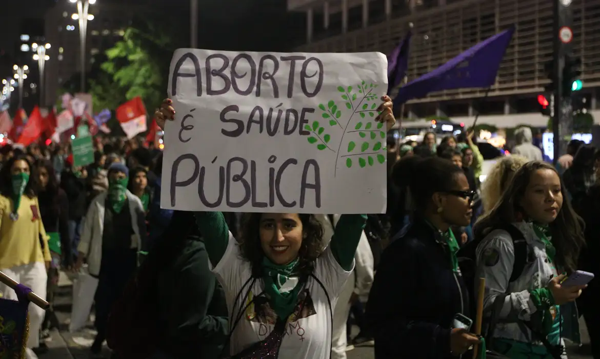 Mulher sorrindo com cartaz dizendo que aborto é questão de saúde pública