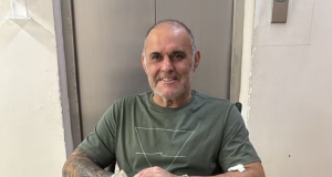 O comerciante Cláudio Crespi sentado em cadeira de rodas, sorrindo, com mala no colo