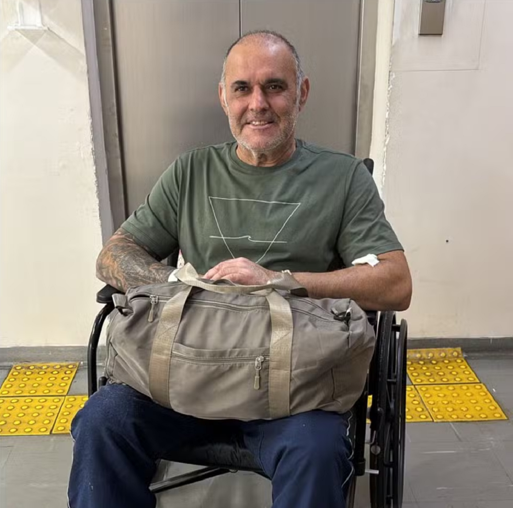 O comerciante Cláudio Crespi sentado em cadeira de rodas, sorrindo, com mala no colo