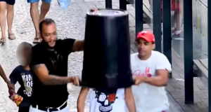 Dois homens colocando um balde na cabeça de outro homem durante pegadinha